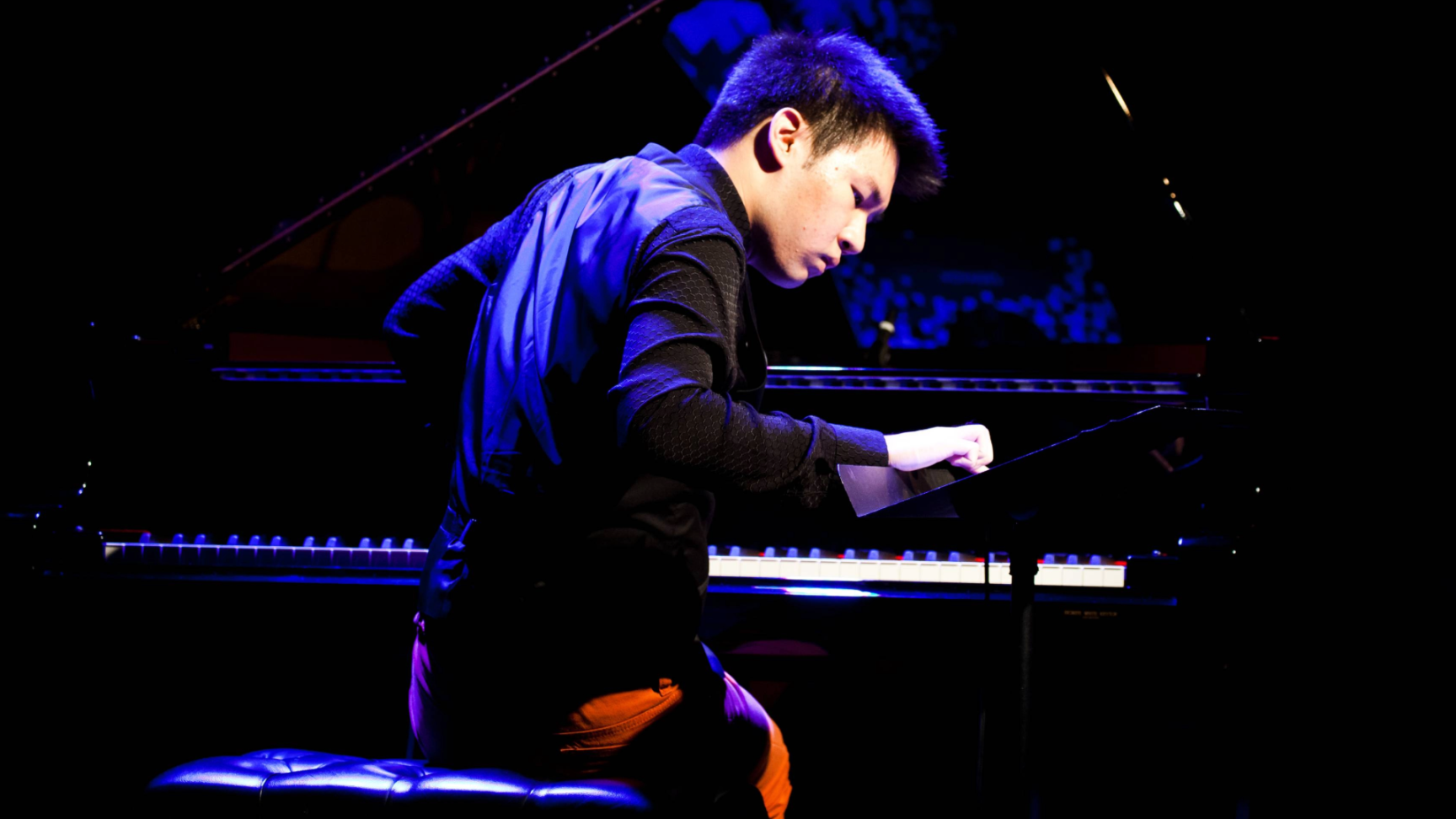 Conrad Tao sitzt auf einer Bühne an einem Flügel und spielt konzentriert Klavier, beleuchtet von blau-violettem Bühnenlicht.