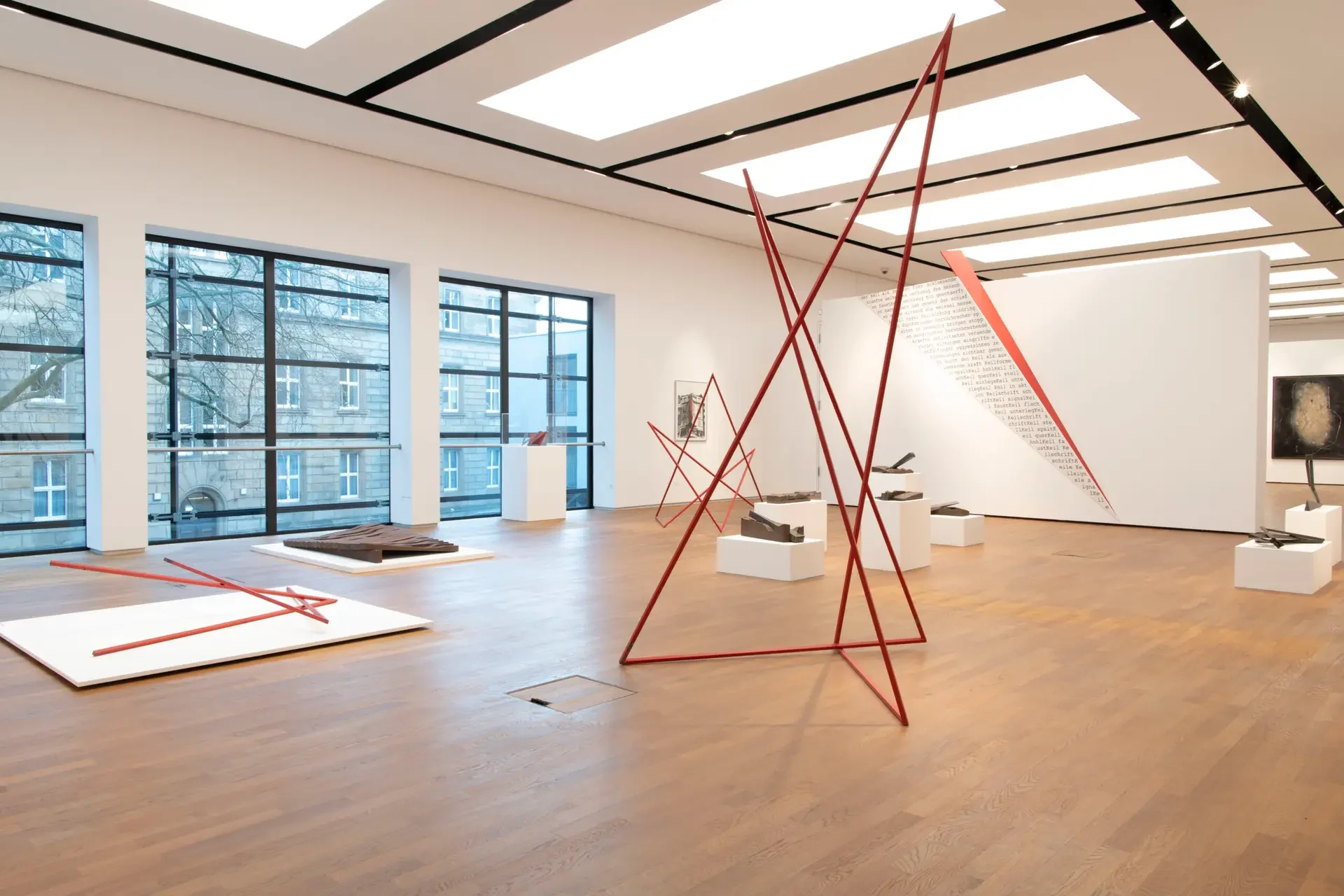 Großer heller Ausstellungsraum mit Holzfußboden und großen Fenstern, darin mehrere abstrakte, rot lackierte Skulpturen aus dünnen Metallstäben von Utz Brocksieper, dazwischen unlackierte Skulpturen aus Stahl, teils auf dem Boden, teils auf weißen Sockeln.