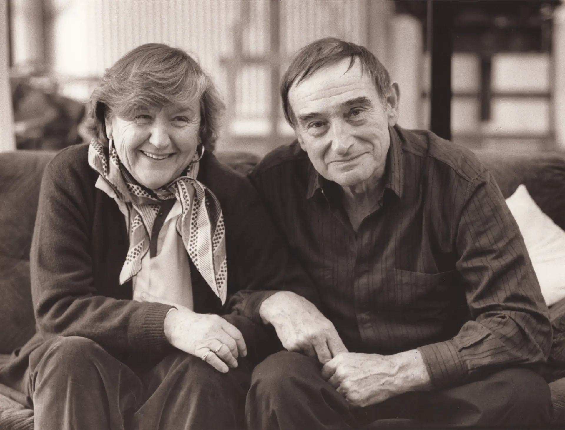 Fotografie in schwarz-weiß: Ulla Schumacher sitzt links mit einem gemusterten Halstuch und dunklem Pullover neben Emil Schumacher, der rechts ein dunkles Hemd trägt, beide sitzen nah beieinander auf einem Sofa.