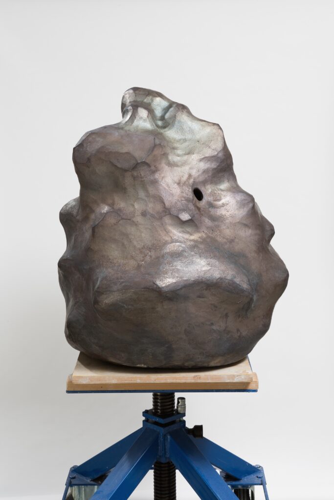 Eine große Skulptur steht zentral auf einem Sockel, der wie ein Drehhocker aussieht. Die Skulptur hat organische Formen und erdige sowie metallische Farben. Sie läuft nach oben hin spitzer zu. Im oberen Teil befindet sich ein Loch.