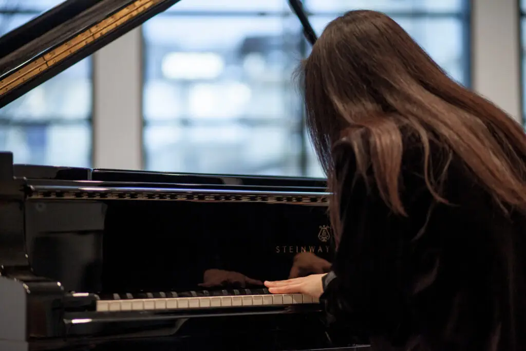 Eine Frau mit langen dunklen Haaren und dunkler Kleidung sitzt an einem schwarzen Steinway-Flügel. Das Bild zeigt die spielenden Hände und Klaviatur sowie den Rücken der spielenden Frau.